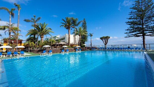Une piscine d’un bleu éclatant, avec des chaises longues et des parasols bleus, entourée de palmiers et d’un bâtiment en arrière-plan.