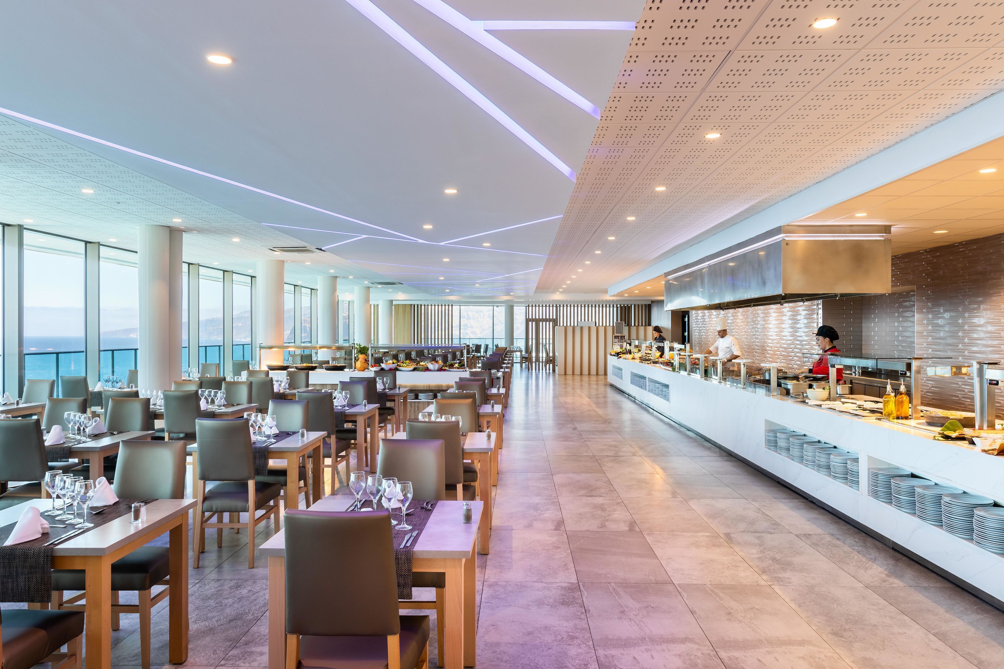 Un restaurante amplio con mesas y sillas largas, una zona de cocina y ventanas grandes que ofrecen vistas.