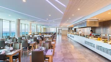 Un restaurante amplio con mesas y sillas largas, una zona de cocina y ventanas grandes que ofrecen vistas.