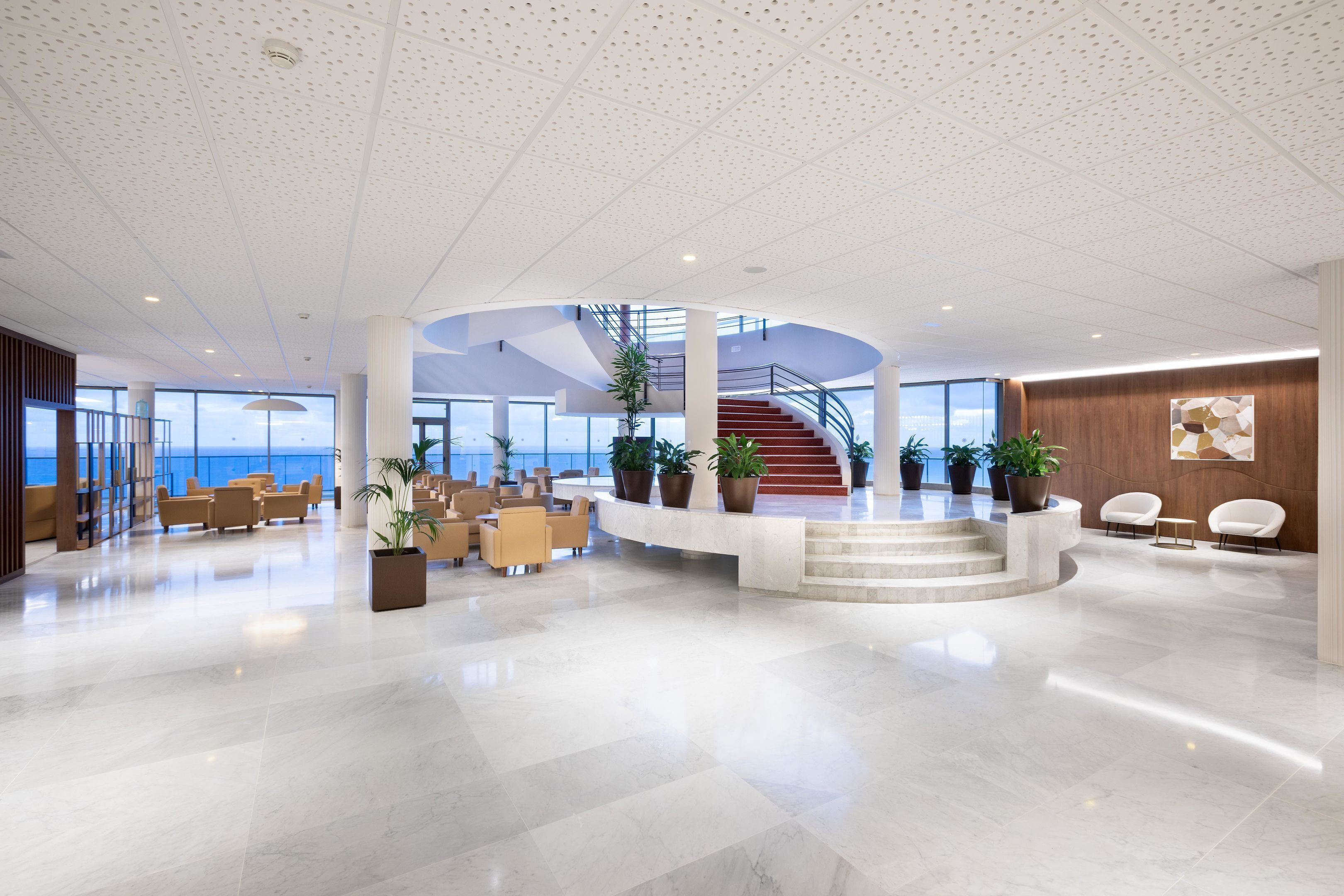 A spacious lobby with a central staircase, seating areas, and potted plants.