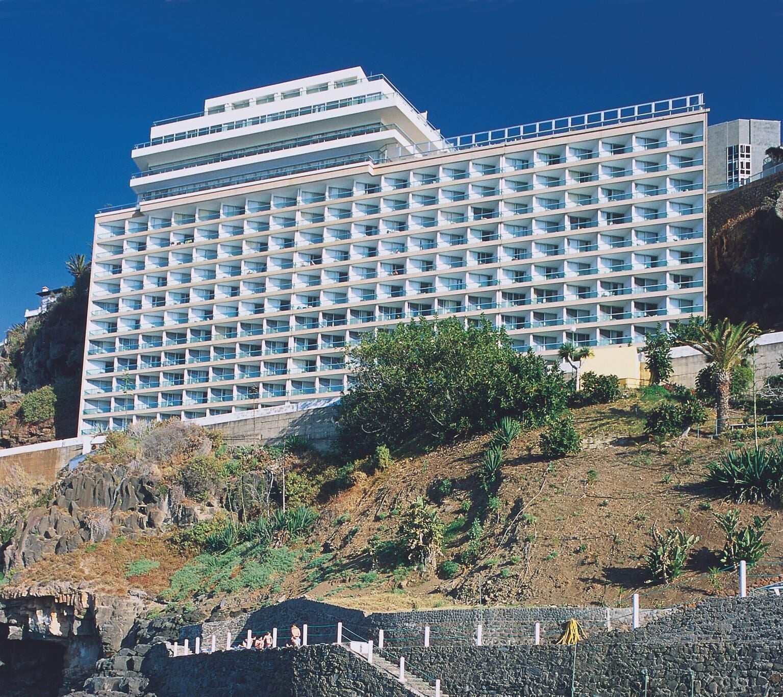 A large, multi-story hotel with a uniform facade, numerous balconies, and a hillside location with sparse vegetation.