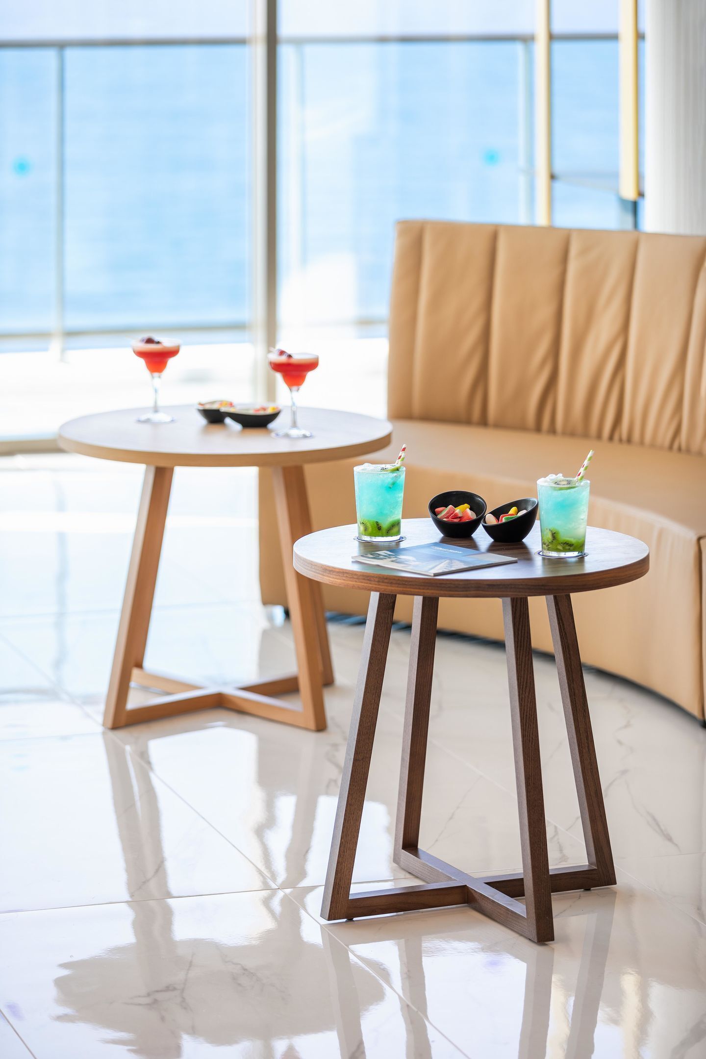 A modern room with a round wooden table and a sofa, both with a light wood finish. There are two glass tables with drinks and bowls on them.