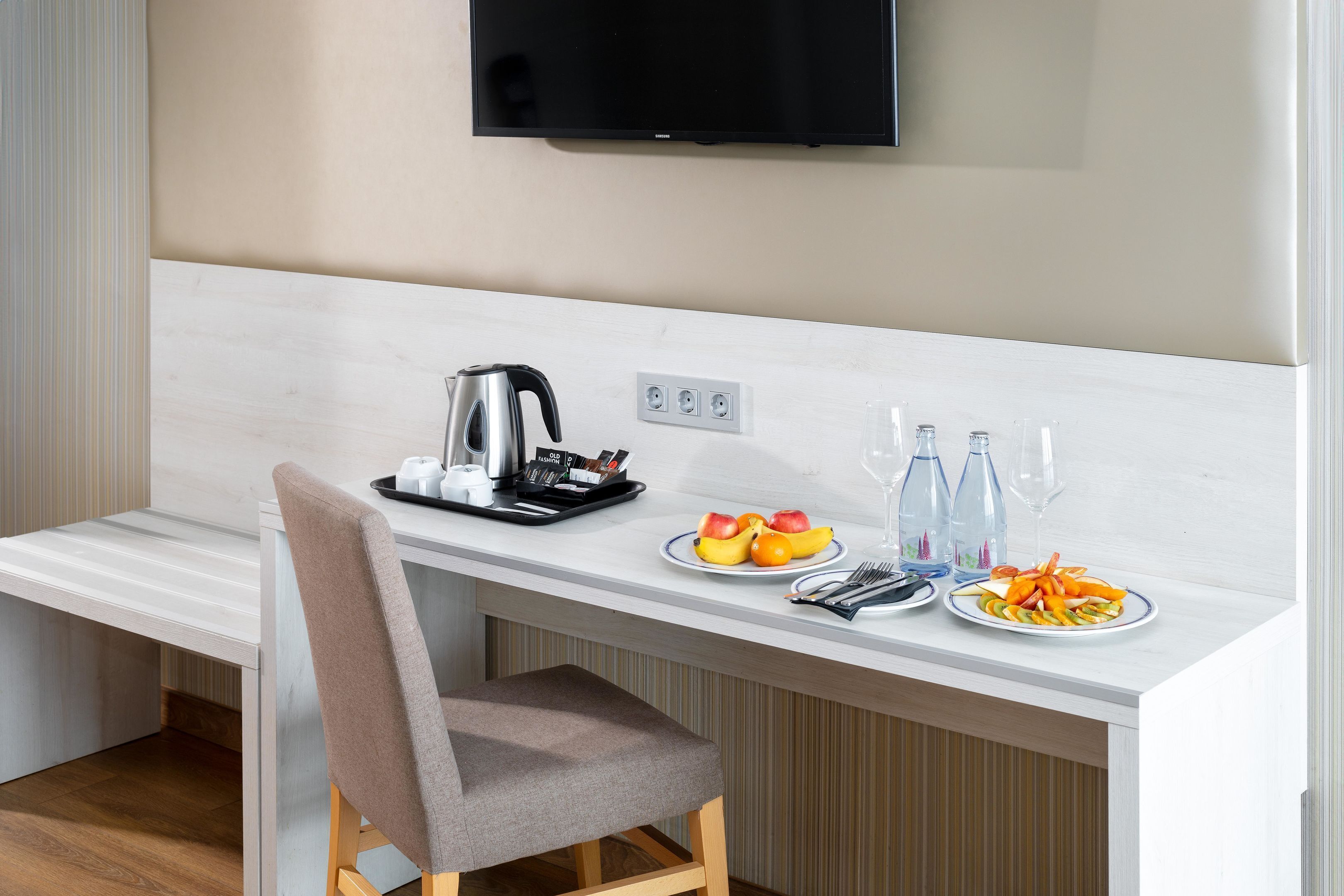 A modern hotel room with a kitchenette, a flat-screen TV mounted on the wall, and a dining area with a chair and a table set with plates of fruit and a kettle.