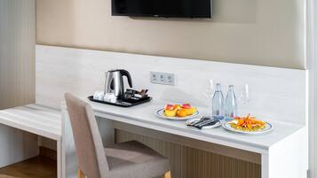 A modern hotel room with a kitchenette, a flat-screen TV mounted on the wall, and a dining area with a chair and a table set with plates of fruit and a kettle.