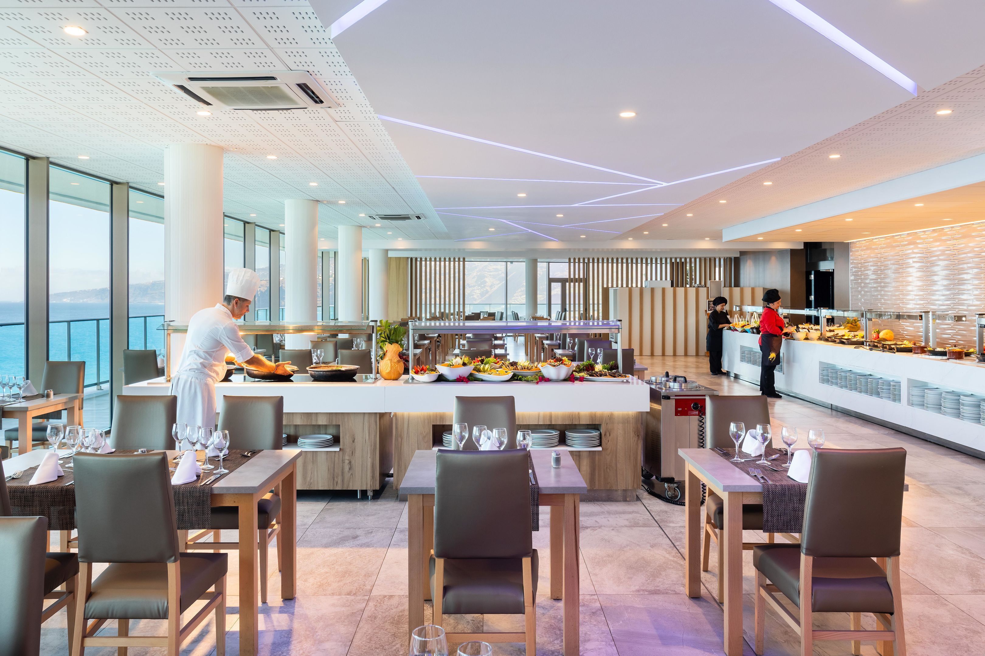 A modern restaurant with a buffet setup, a chef preparing food, and a view of the sea.