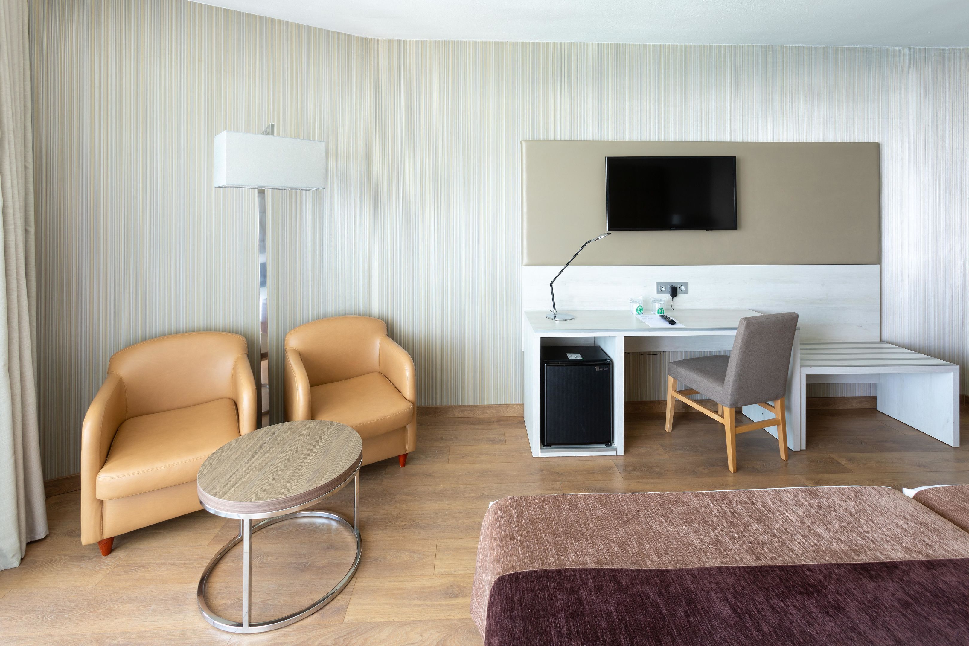 A hotel room with a bed, two armchairs, a desk with a computer, a TV, and a lamp.