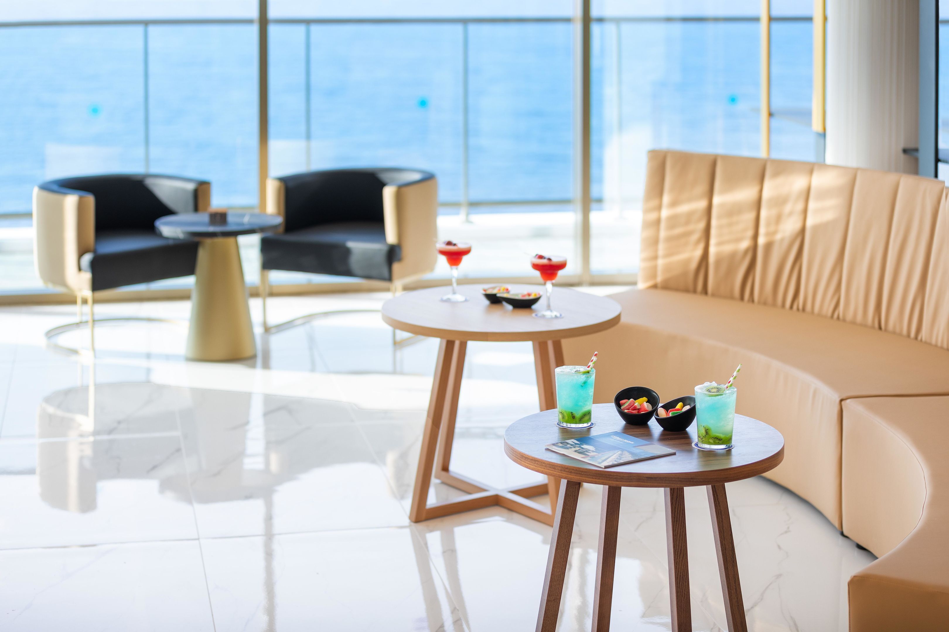 A modern lounge area with a sofa, two armchairs, and a round table with drinks and a bowl of fruit.