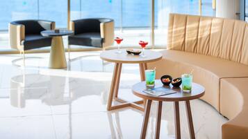 A modern lounge area with a sofa, two armchairs, and a round table with drinks and a bowl of fruit.