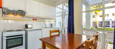 Eine kompakte Küche mit weißen Schränken, einem Herd und einem kleinen Kühlschrank. Ein Holztisch mit Stühlen und ein großes Fenster bieten Blick ins Freie.