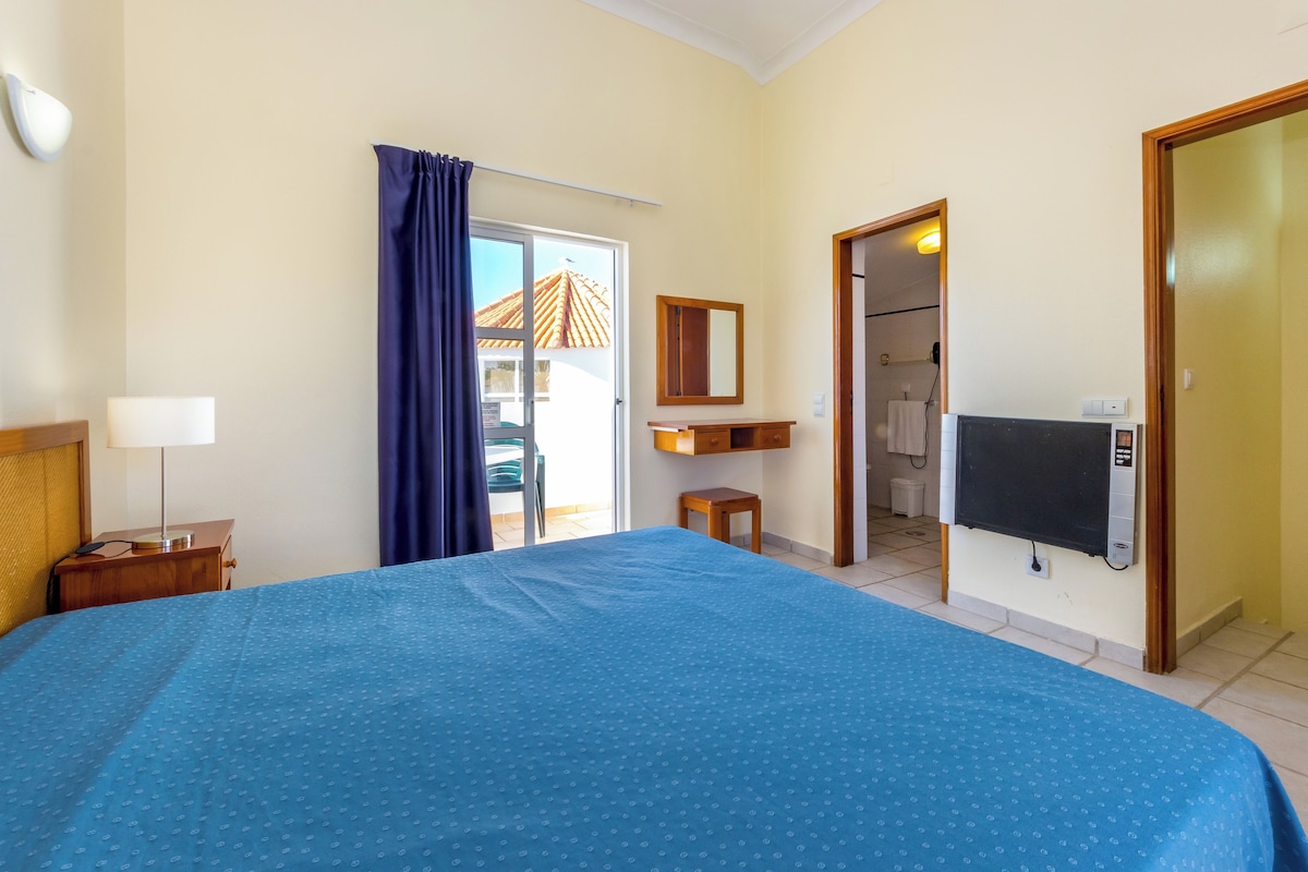 A hotel room with a large bed, a desk with a lamp, a TV, and a bathroom visible through an open door.