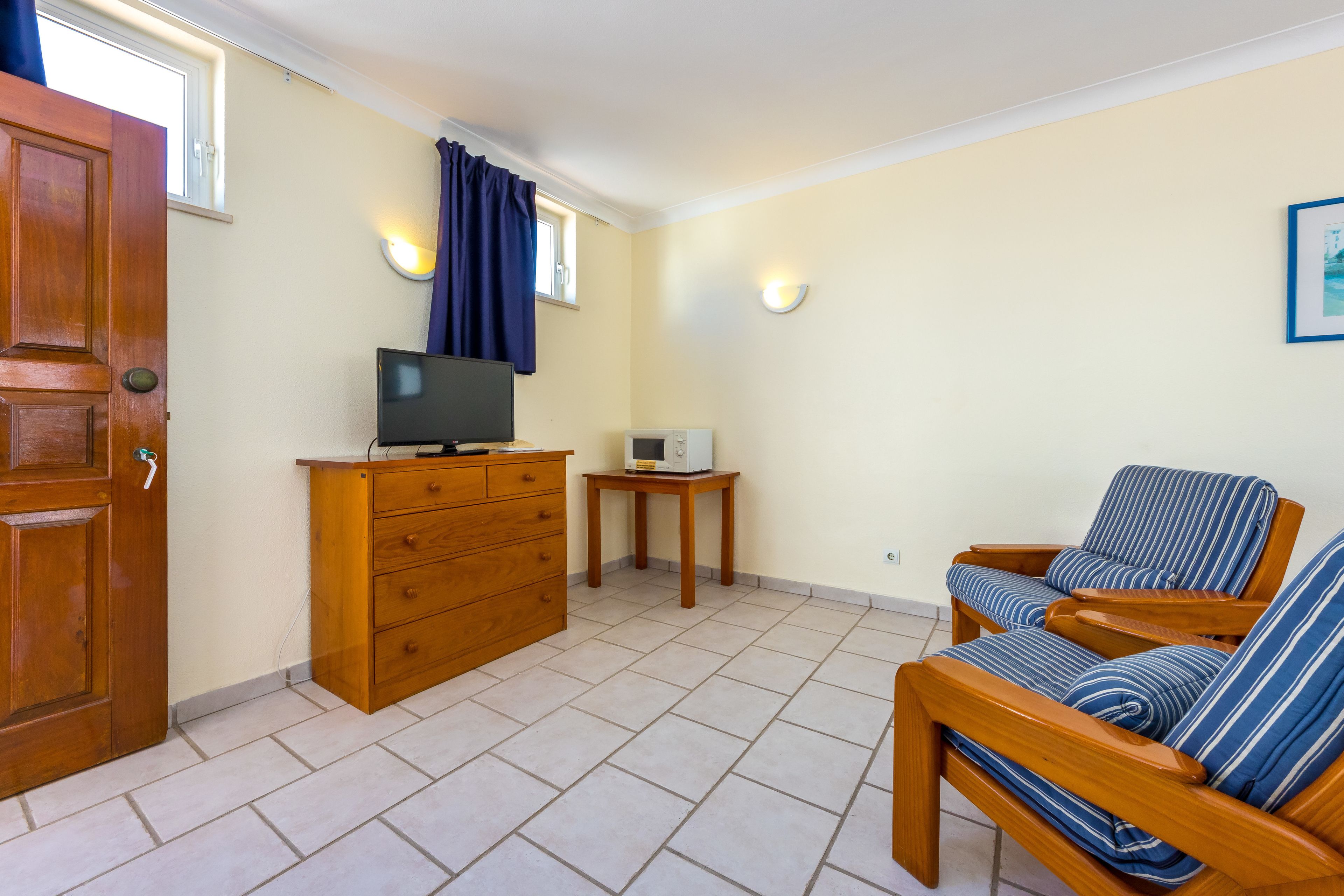 Een kamer met een houten deur, een televisie op een dressoir, een klein tafeltje met een magnetron en een gestreepte stoel.