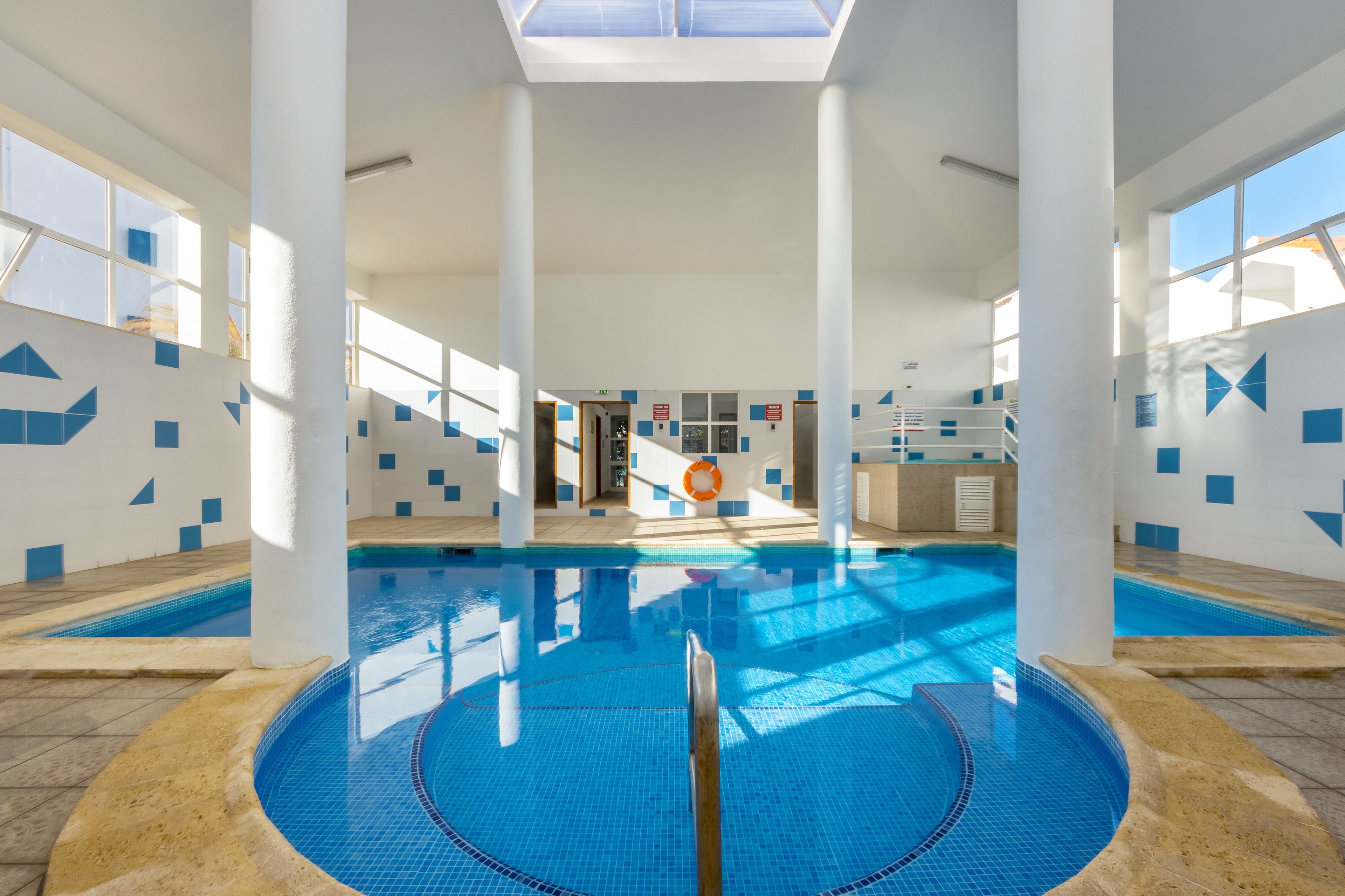 Une piscine intérieure à l’eau d’un bleu cristallin, entourée de murs blancs ornés de motifs géométriques bleus, et dotée de grandes fenêtres laissant entrer la lumière naturelle.