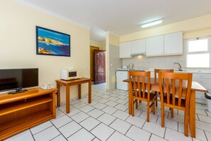 A compact kitchen with white cabinets, a microwave, a toaster, and a wooden dining table with chairs. A television is on a wooden stand, and a painting of a coastal scene is on the wall.