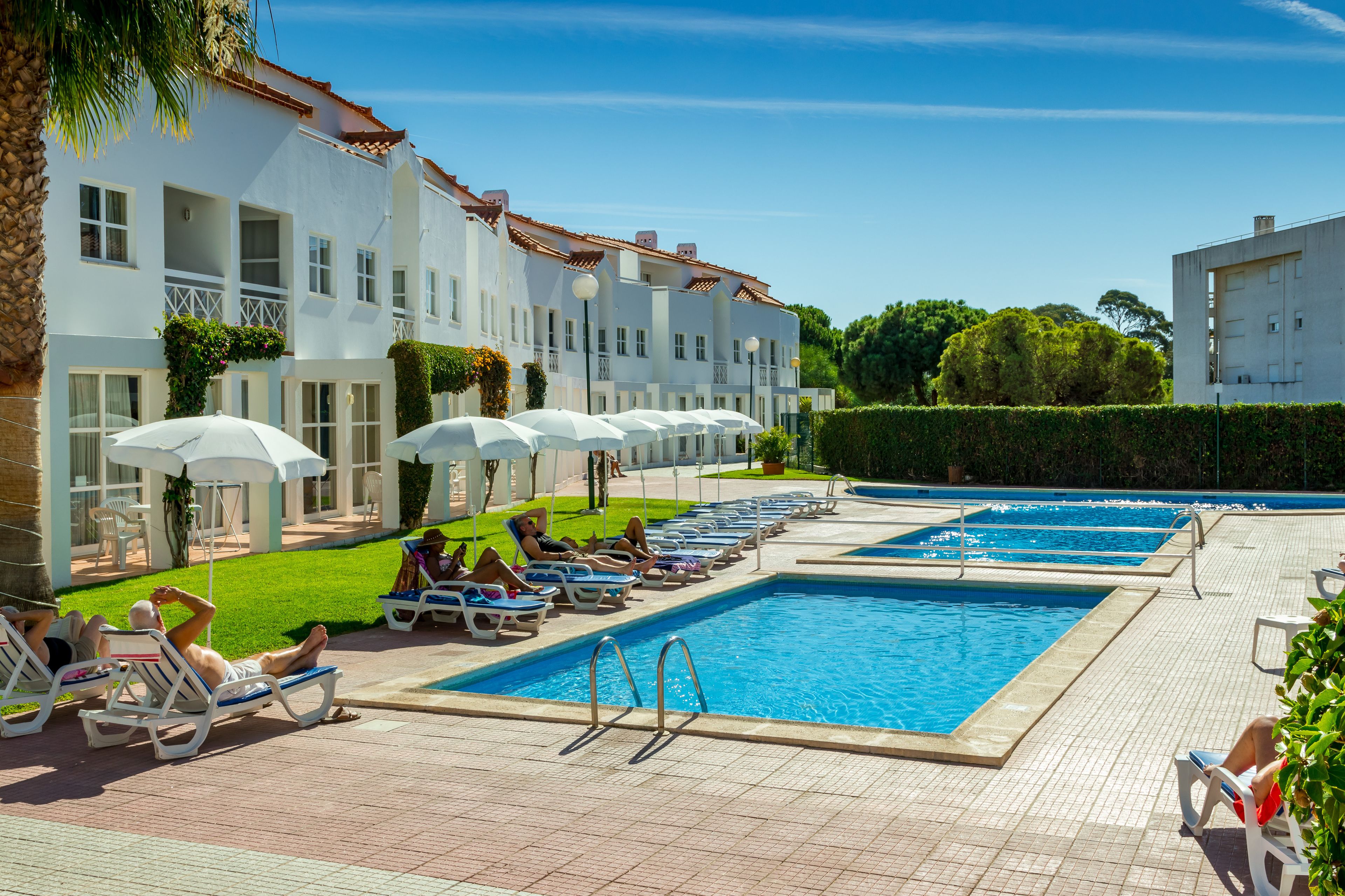 A pool area with lounge chairs and umbrellas, a clear blue sky, and a building with balconies.