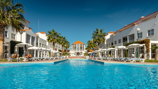 A swimming pool with lounge chairs and umbrellas, surrounded by white buildings with red roofs and palm trees. - Apartamentos Turísticos Club Ouratlântico (Albufeira)