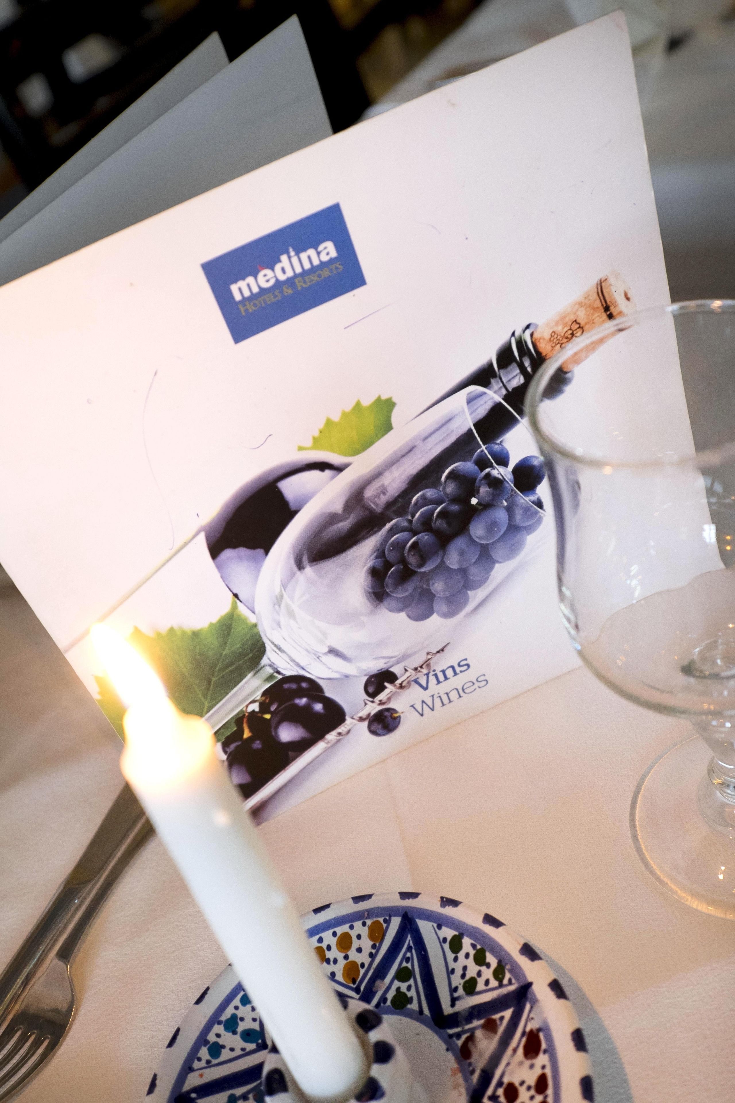 Una mesa servida con un vaso de agua, una vela, un plato con un diseño azul y blanco, y un folleto de Medina Hotels & Resorts presentando Vins Wines.