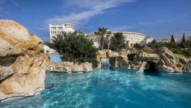 A clear pool surrounded by rocky formations and palm trees, with a large building in the background. - Medina Solaria & Thalasso (Hammamet)