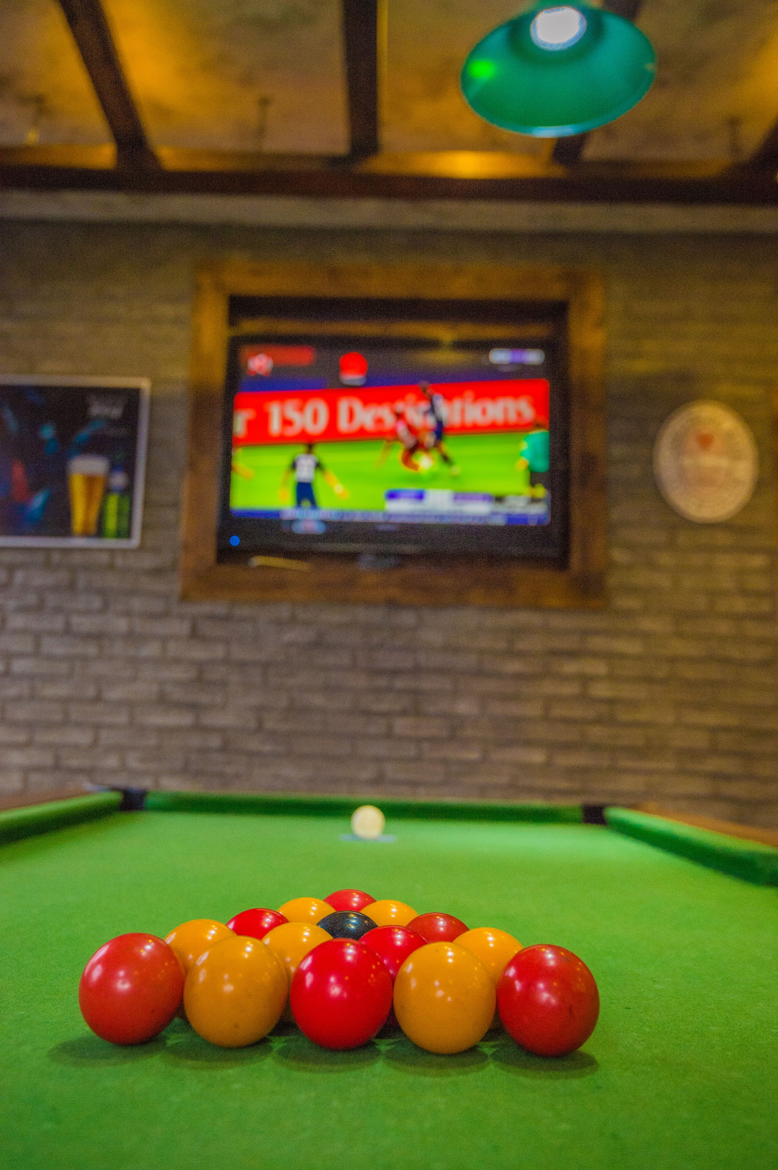 A pool table with balls arranged in a triangle, a television displaying a sports game, and a brick wall in the background.