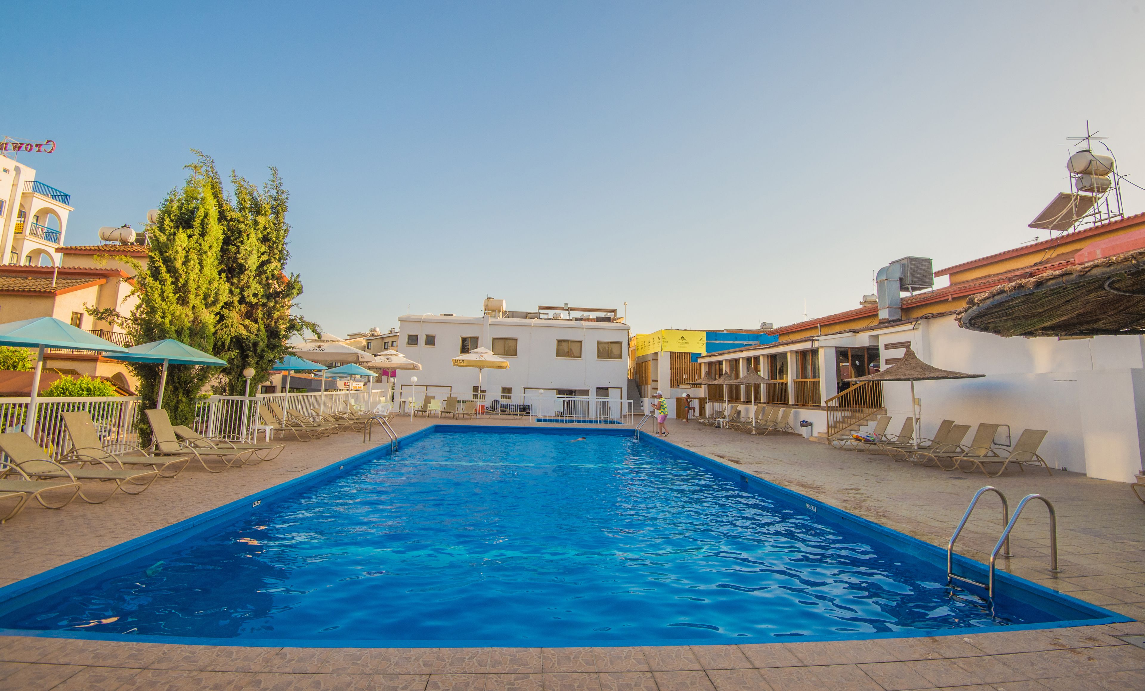 Una piscina con sillas de descanso y sombrillas, rodeada de edificios y árboles.