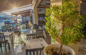 An outdoor dining area with tables and chairs, a potted tree, and a view of the street. - Windmills Hotel Apartments (Paralimni)