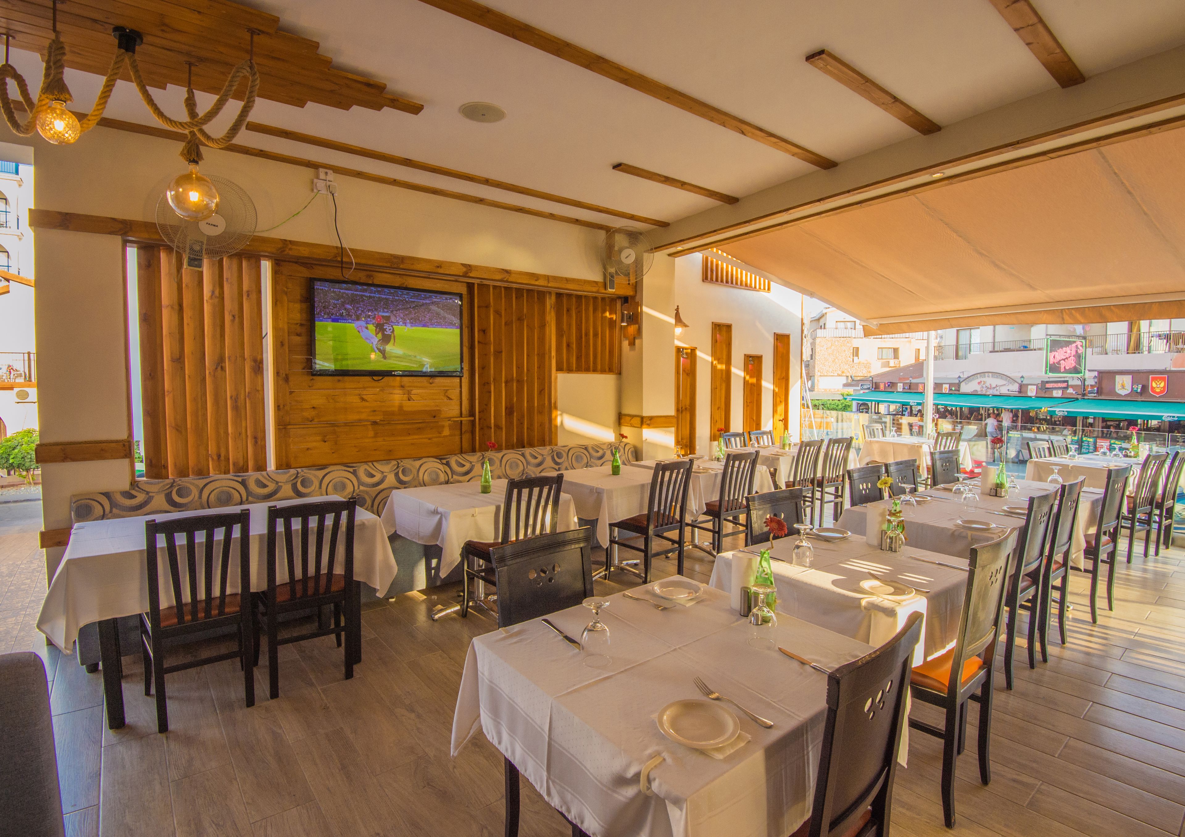 An outdoor restaurant with wooden beams, tables set with white tablecloths, and a TV screen displaying a sports event.