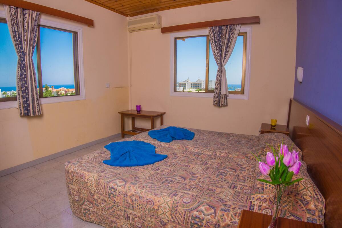 Un dormitorio con una colcha estampada, dos ventanas con cortinas, un cabecero de madera, una mesita de noche pequeña con una lámpara y un jarrón con flores.
