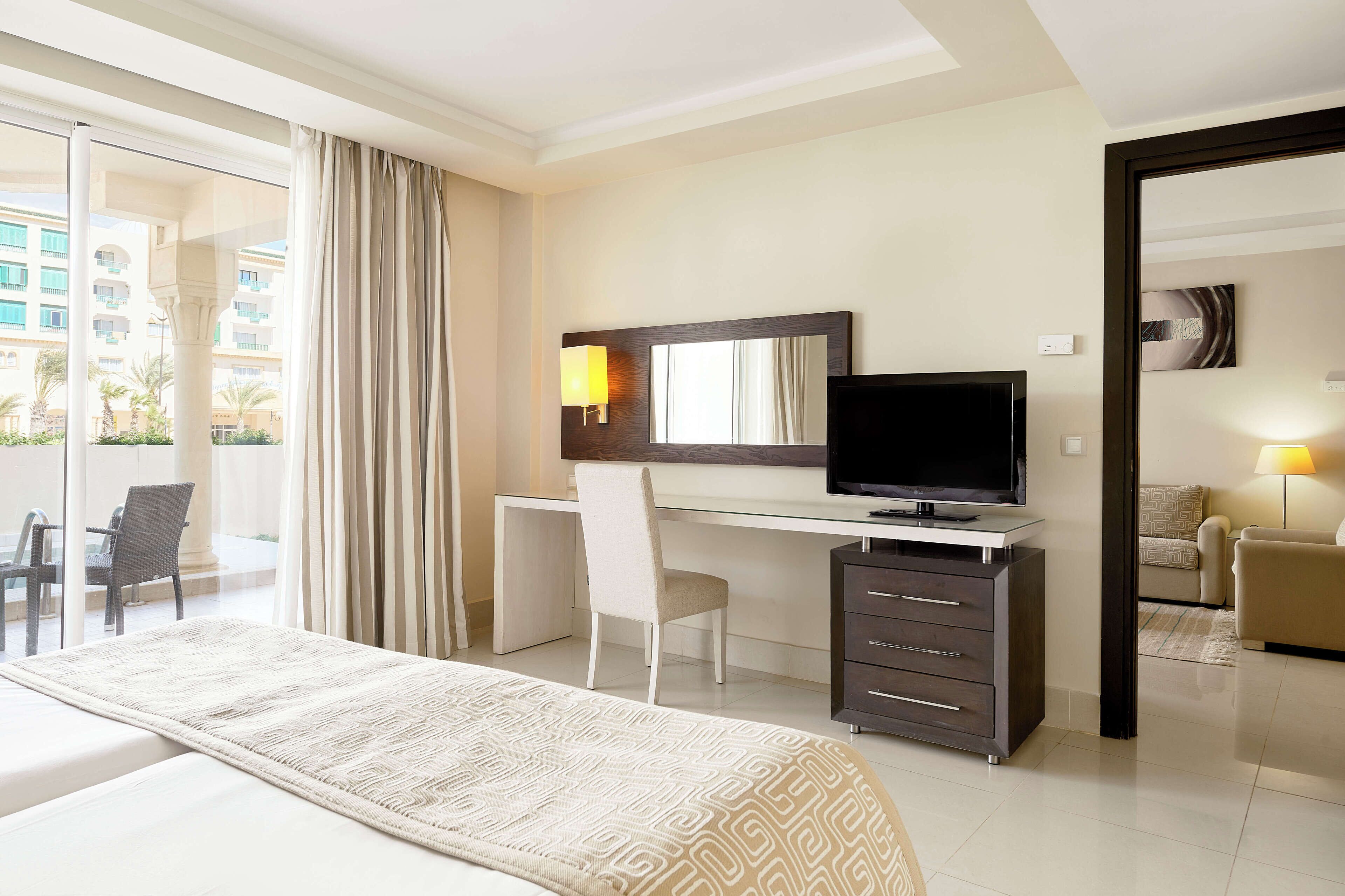 A hotel room with a bed, a desk with a TV, a chair, a dresser, and a balcony with a view of buildings.