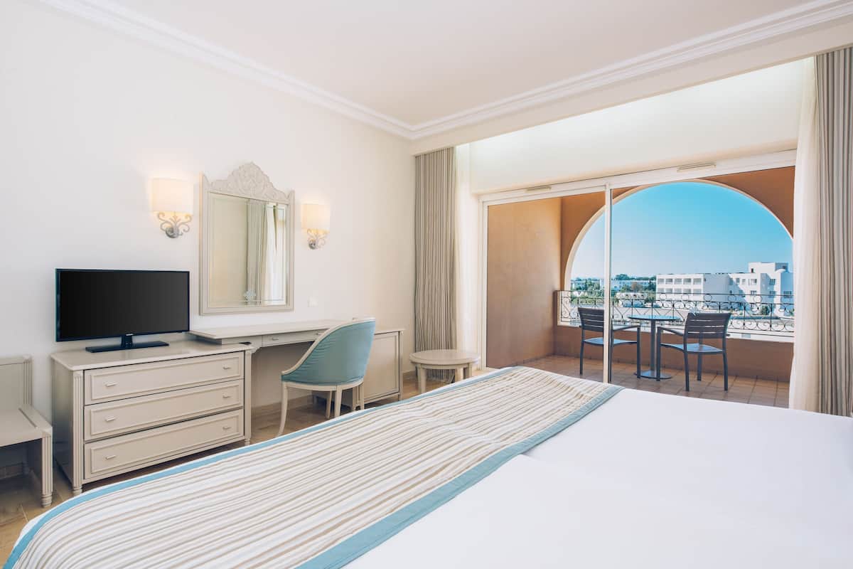 Een hotelkamer met een bed, een bureau met een televisie, een stoel, een klein tafeltje en een groot raam met uitzicht op gebouwen.