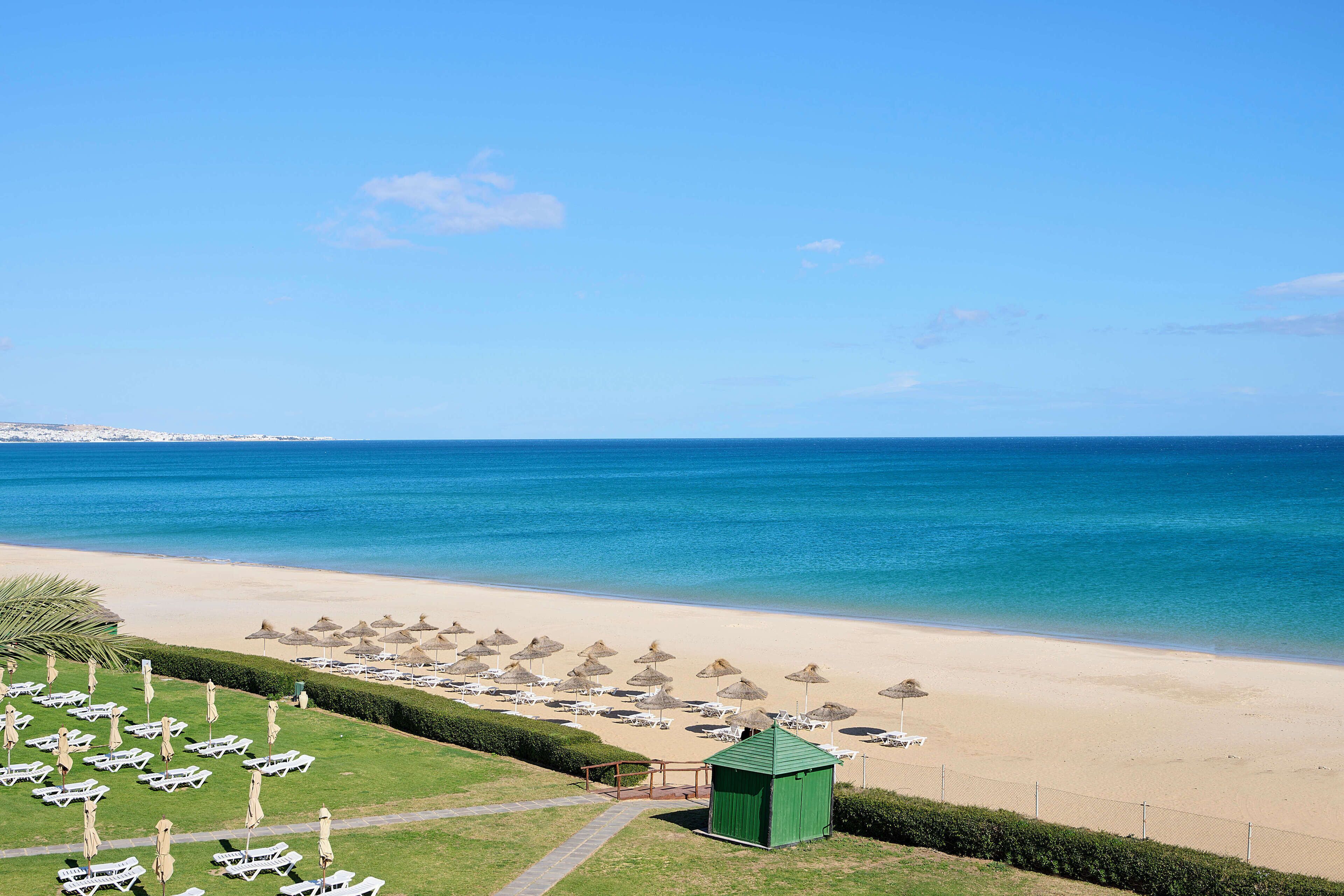 A beach with rows of sun loungers and umbrellas, a small green structure, and a clear view of the ocean.