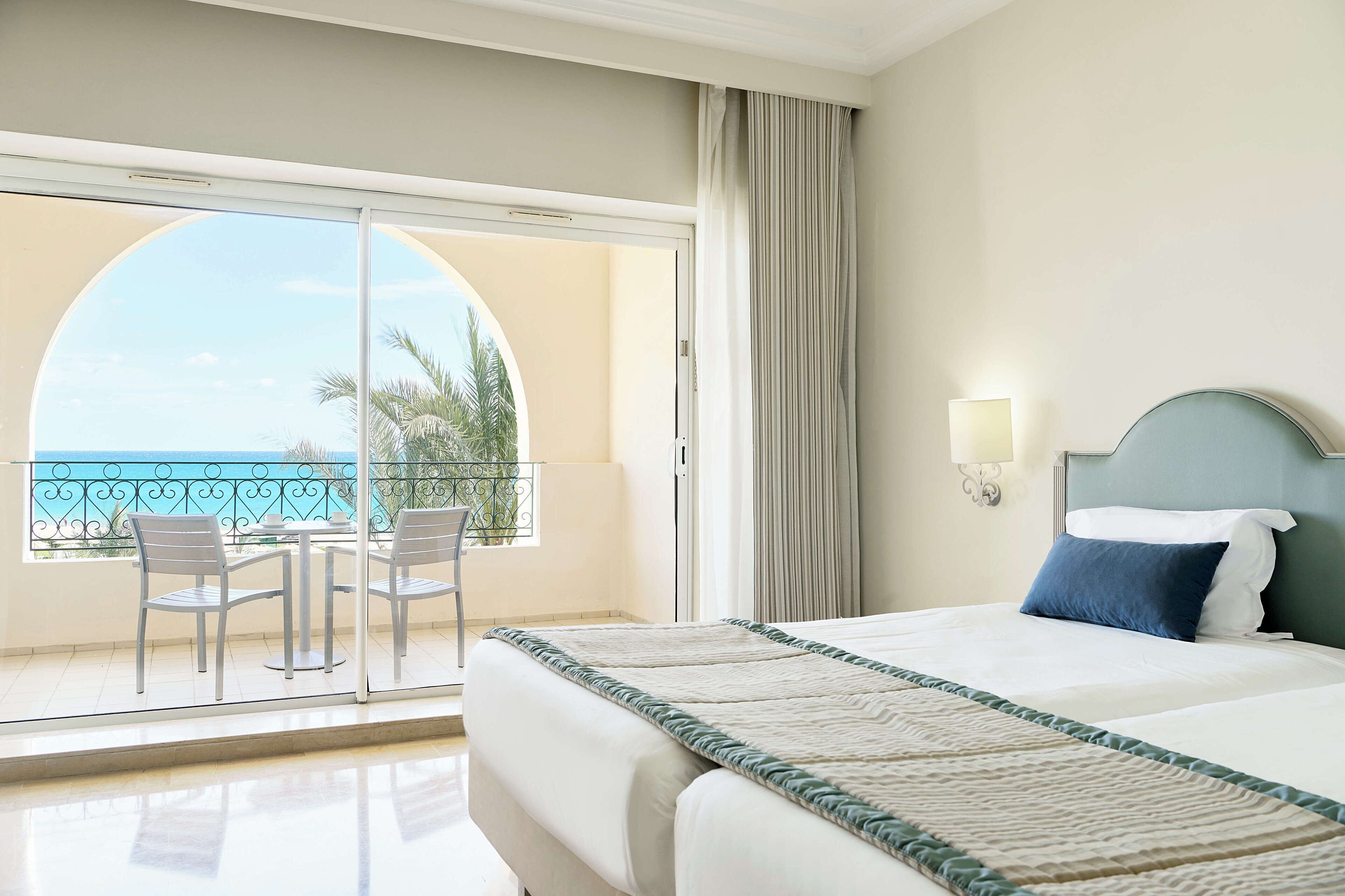 A hotel room with a bed, a view of the ocean, a balcony with chairs, and a large window.