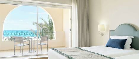 A hotel room with a bed, a view of the ocean, a balcony with chairs, and a large window.
