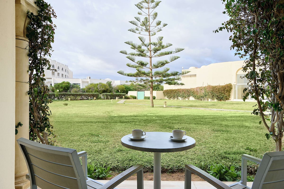 Een terras met twee stoelen en een tafel, een kleine boom en een gazon met een gebouw op de achtergrond.