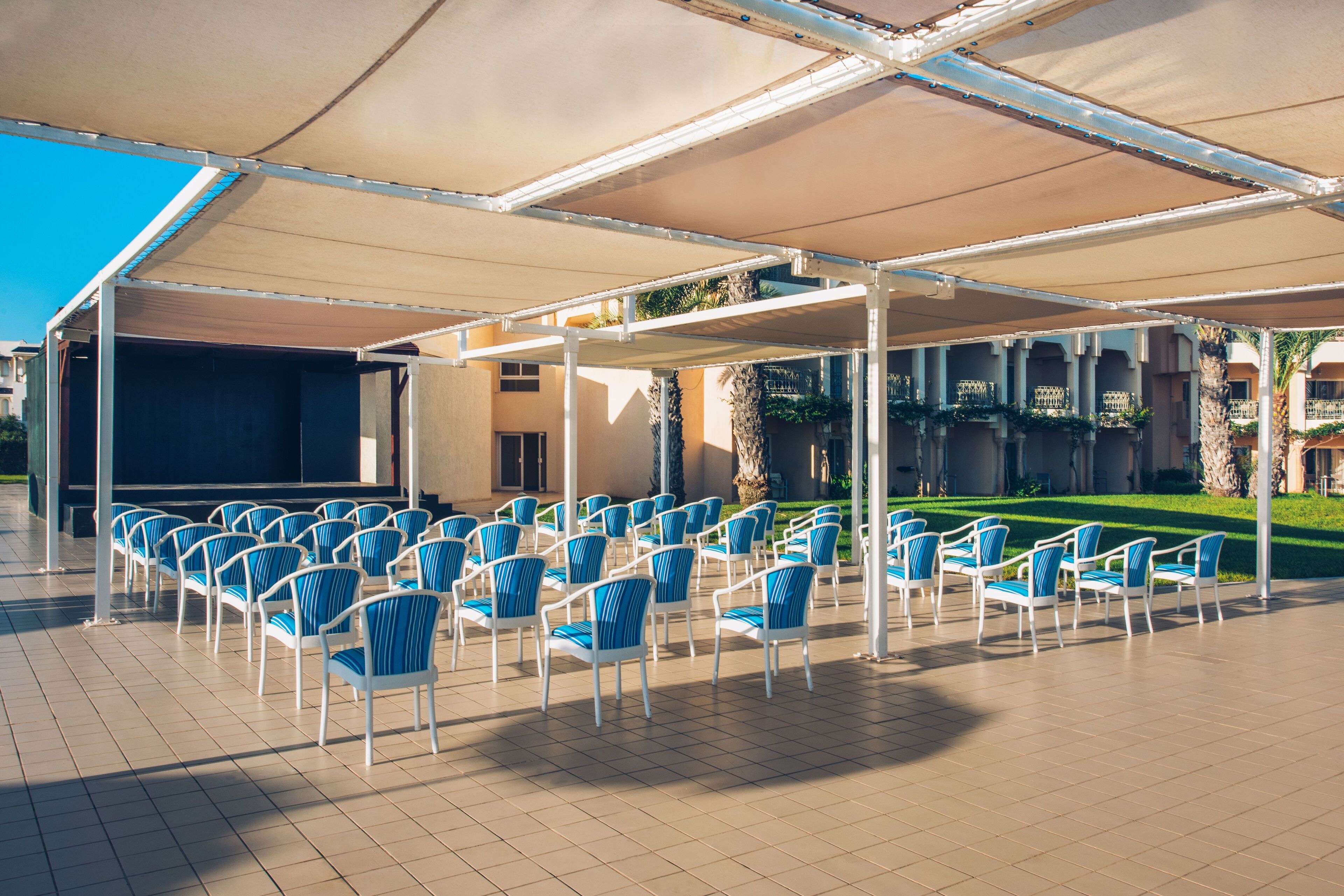 Un espacio al aire libre con filas de sillas bajo una pérgola, un escenario y vistas a un edificio con columnas y un césped.