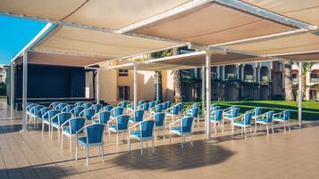Un espacio al aire libre con filas de sillas bajo una pérgola, un escenario y vistas a un edificio con columnas y un césped.