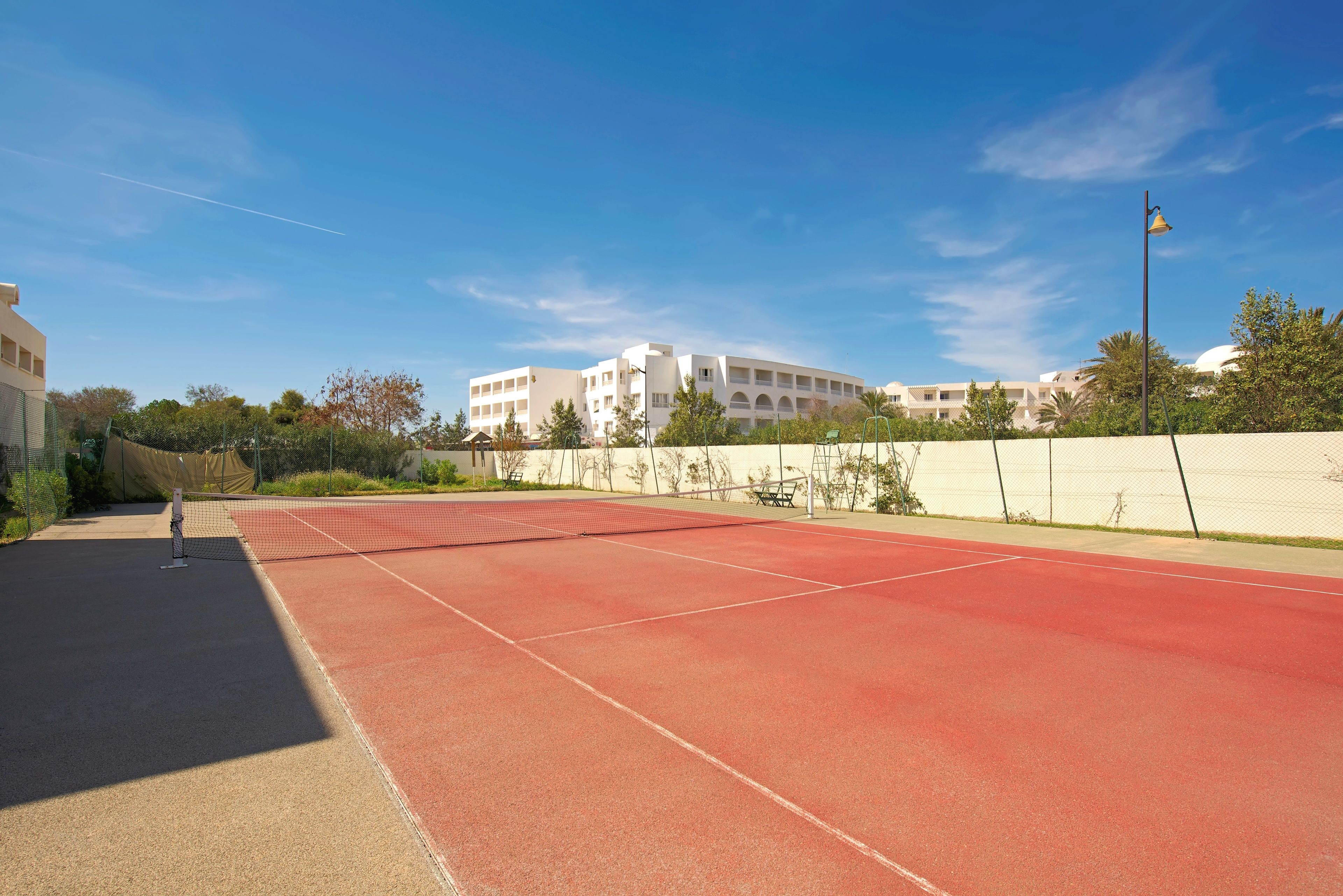 Una cancha de tenis al aire libre con una red, rodeada por una valla y edificios en el fondo.