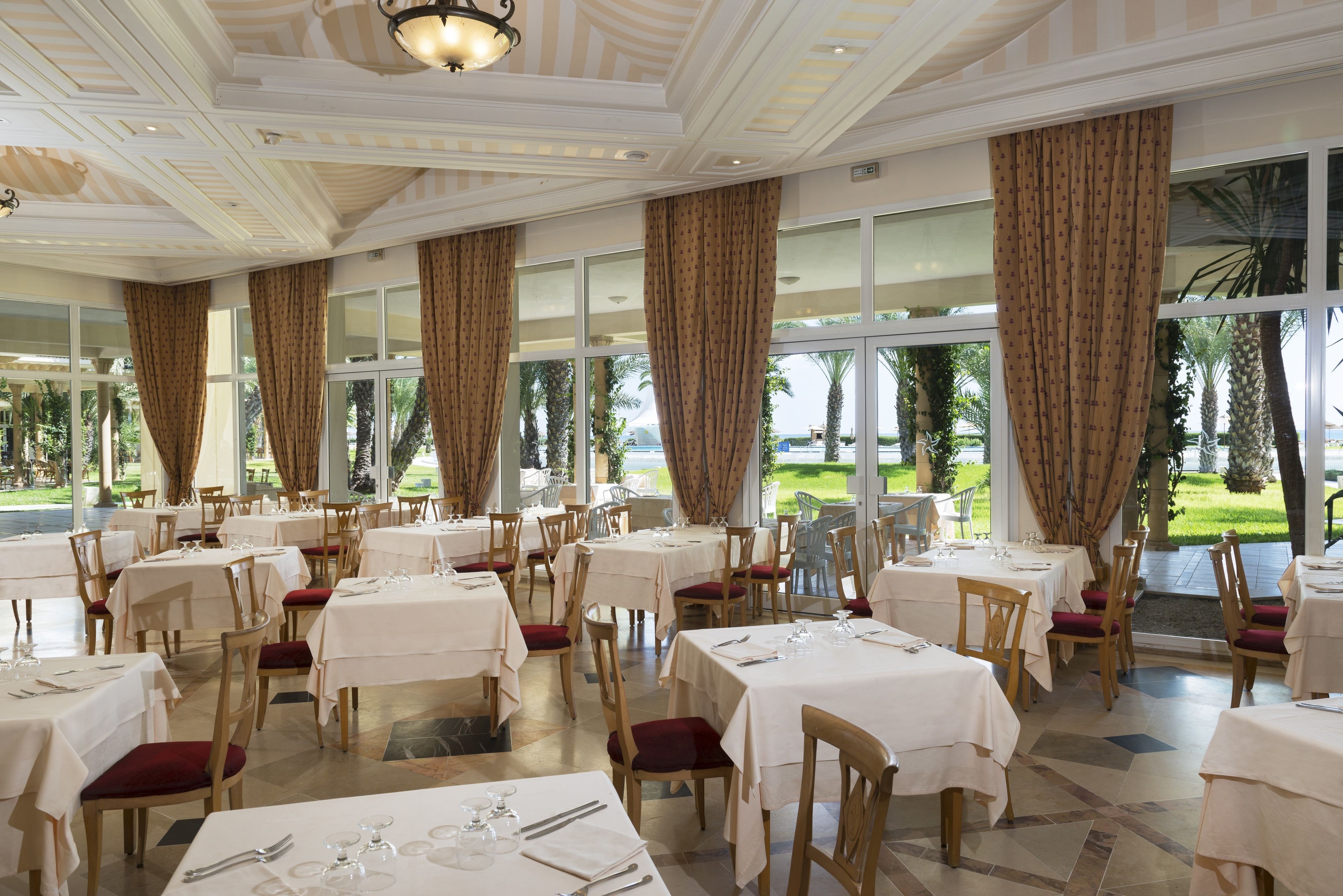 A restaurant with multiple tables and chairs, large windows with curtains, and a view of a landscaped garden.