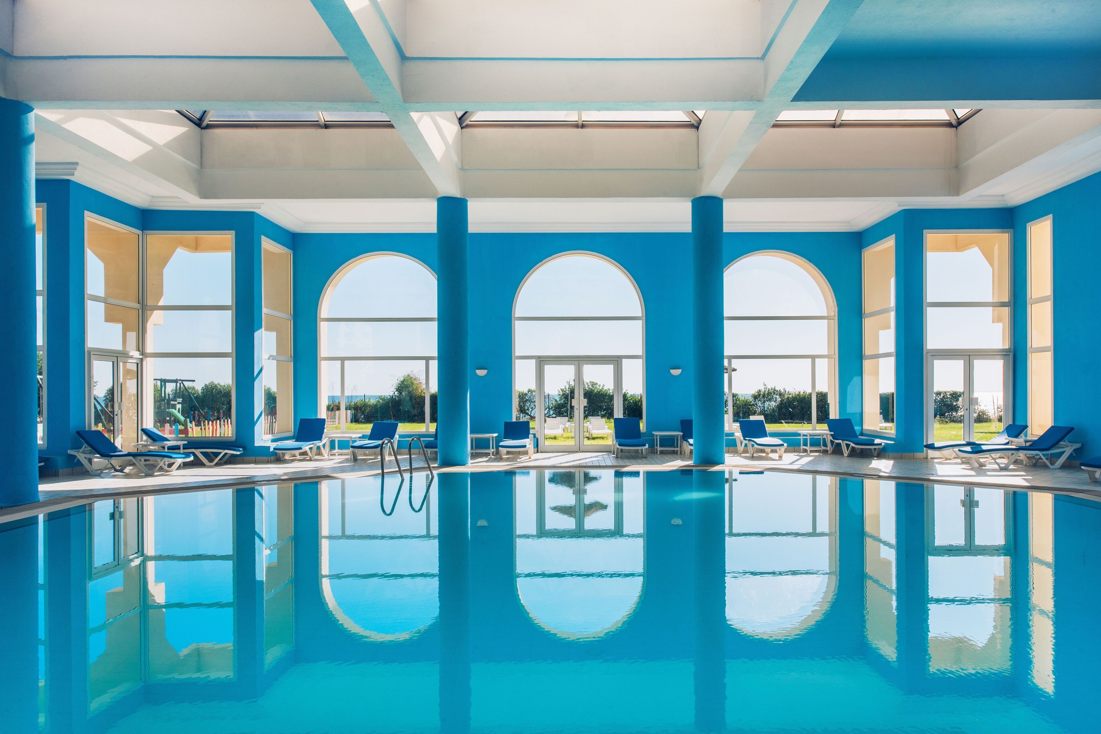Una gran piscina interior amb aigua cristal·lina blava, envoltada de parets blaves i grans finestres d'arc.