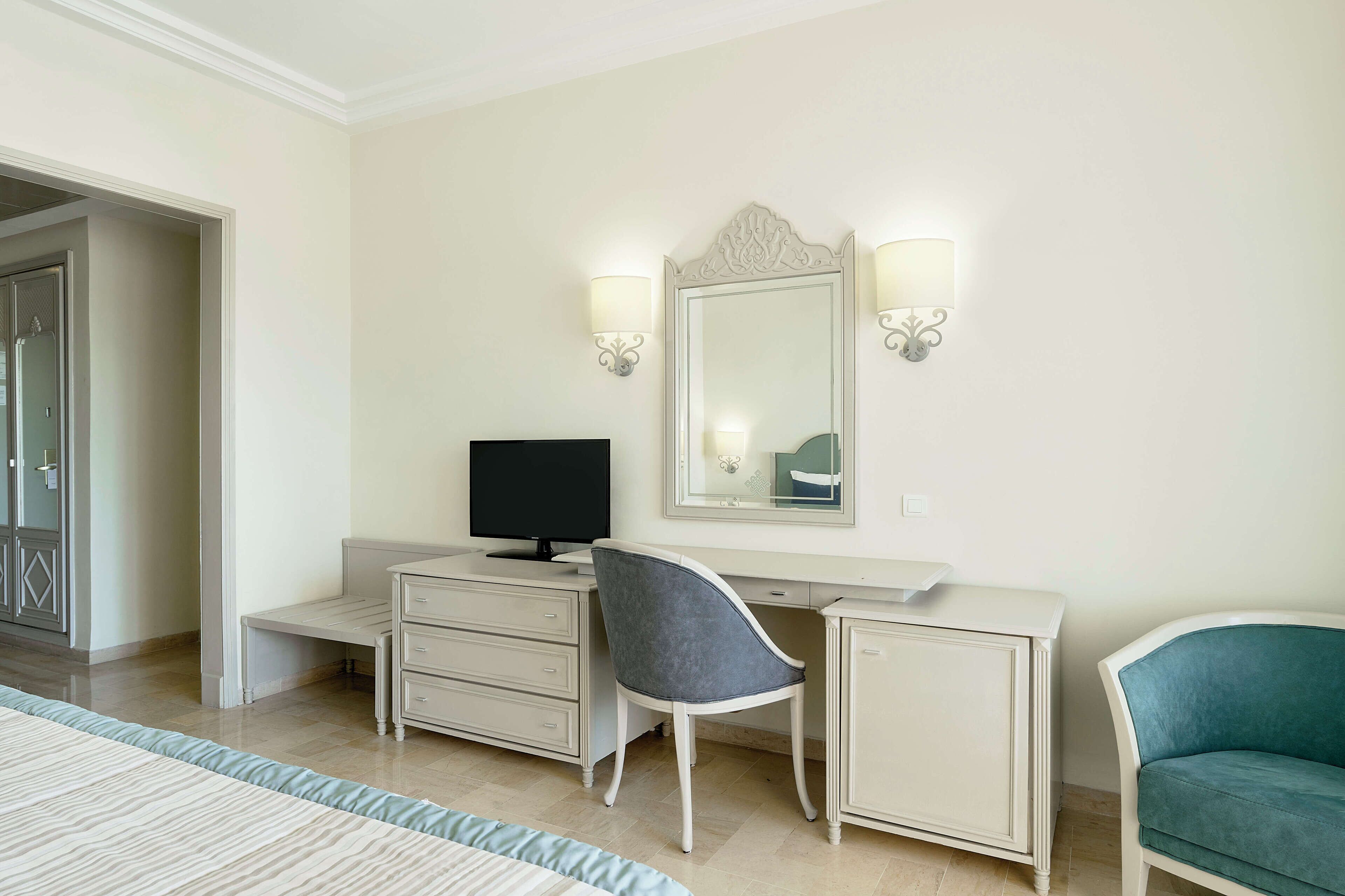 A hotel room with a bed, a desk with a computer, a chair, a dresser, and a mirror.