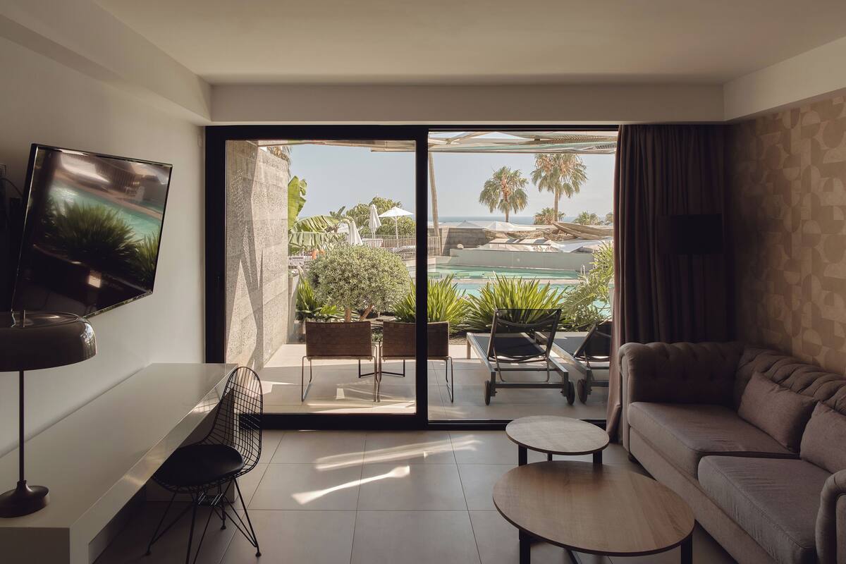 A modern living room with a large TV, a sofa, a coffee table, and a balcony with a pool view.