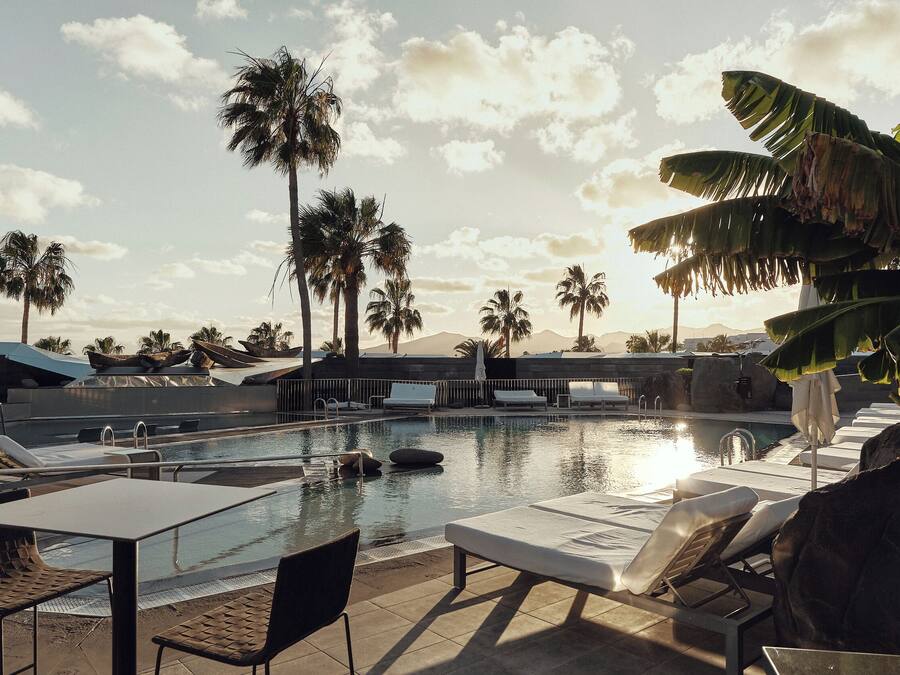 A pool area with lounge chairs and tables, surrounded by palm trees, under a partly cloudy sky.