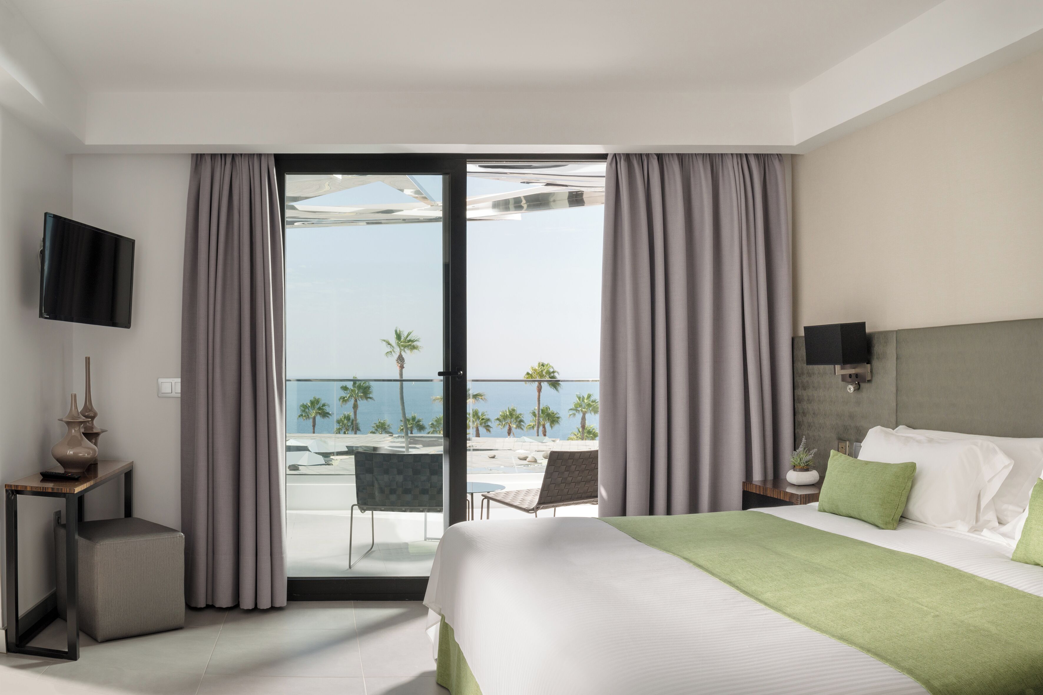 A hotel room with a bed, a TV, a balcony with a view of the sea, and a small table with a vase.