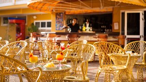Outdoor wicker chairs and tables with drinks, a bar area, and a yellow building facade. - Caleta Garden (Antigua)
