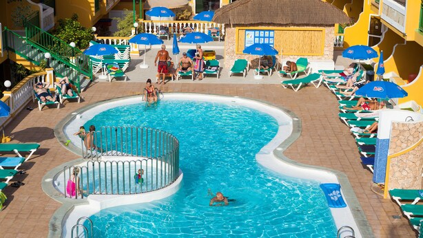 A swimming pool with sun loungers and umbrellas, surrounded by a tiled deck and a building with a thatched roof. - Caleta Garden (Antigua)