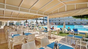 A covered outdoor area with tables and chairs, a pool, and a view of a building in the background. - Hotel Apartamento Brisa Sol (Albufeira)