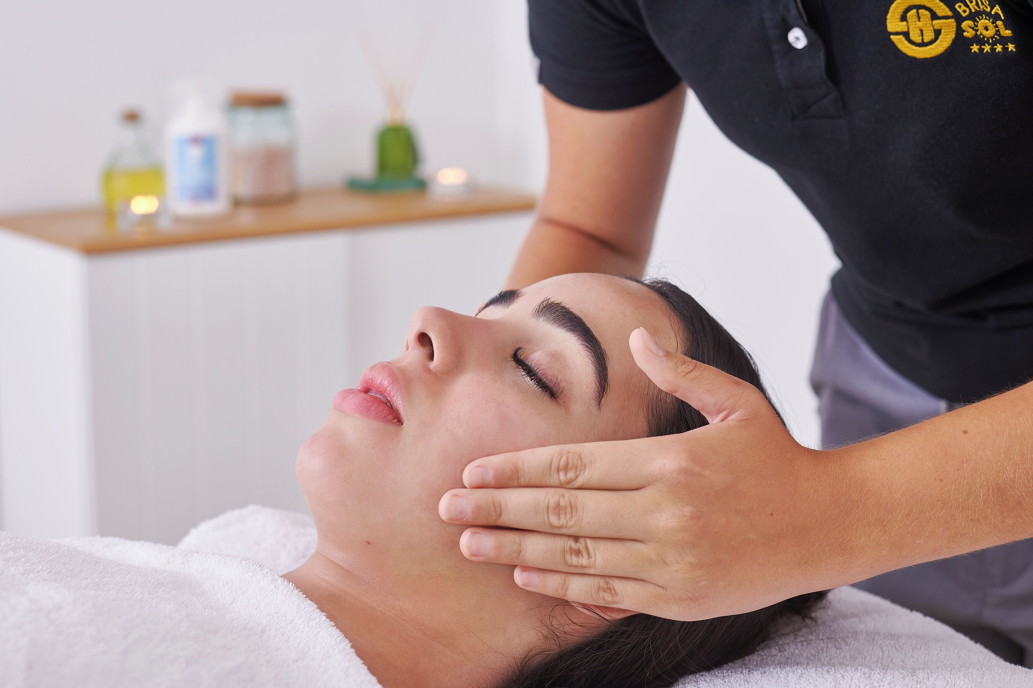 A person receiving a facial treatment in a room with a white wall and a wooden shelf with various items.