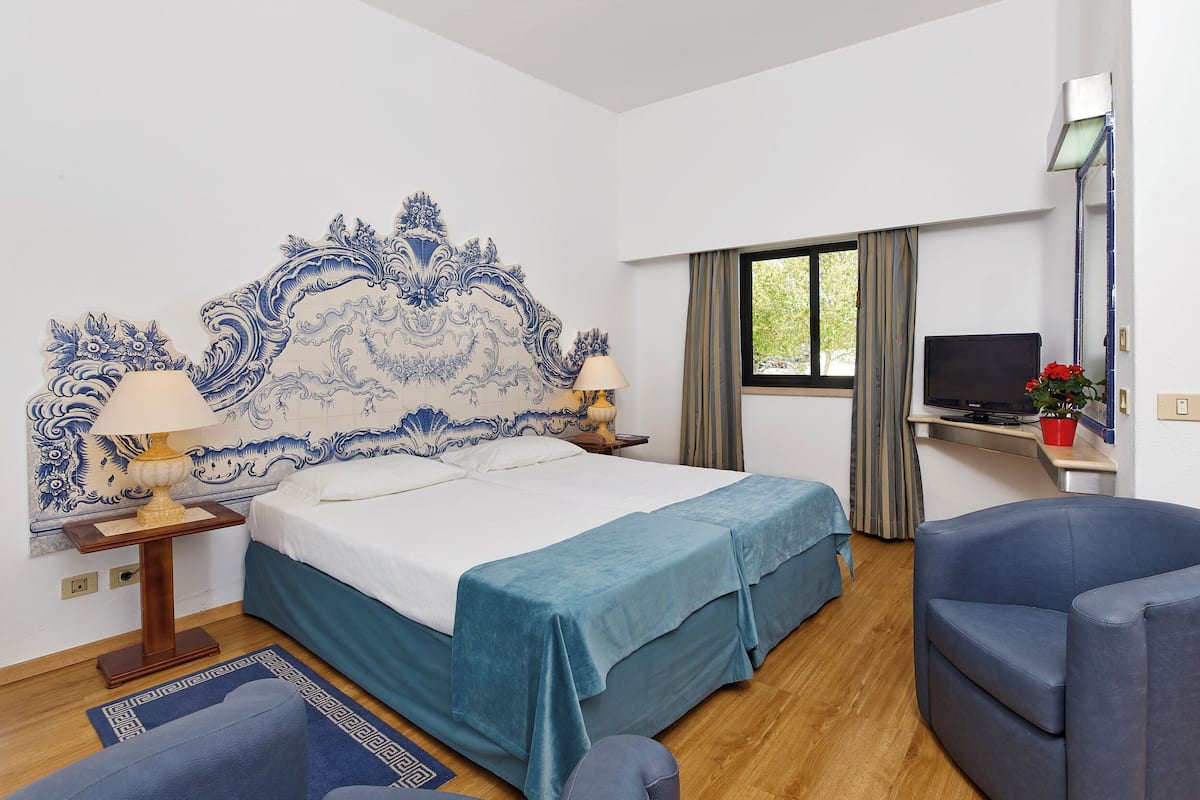 Quarto de hotel com cama, escrivaninha, cadeira, televisão e janela.