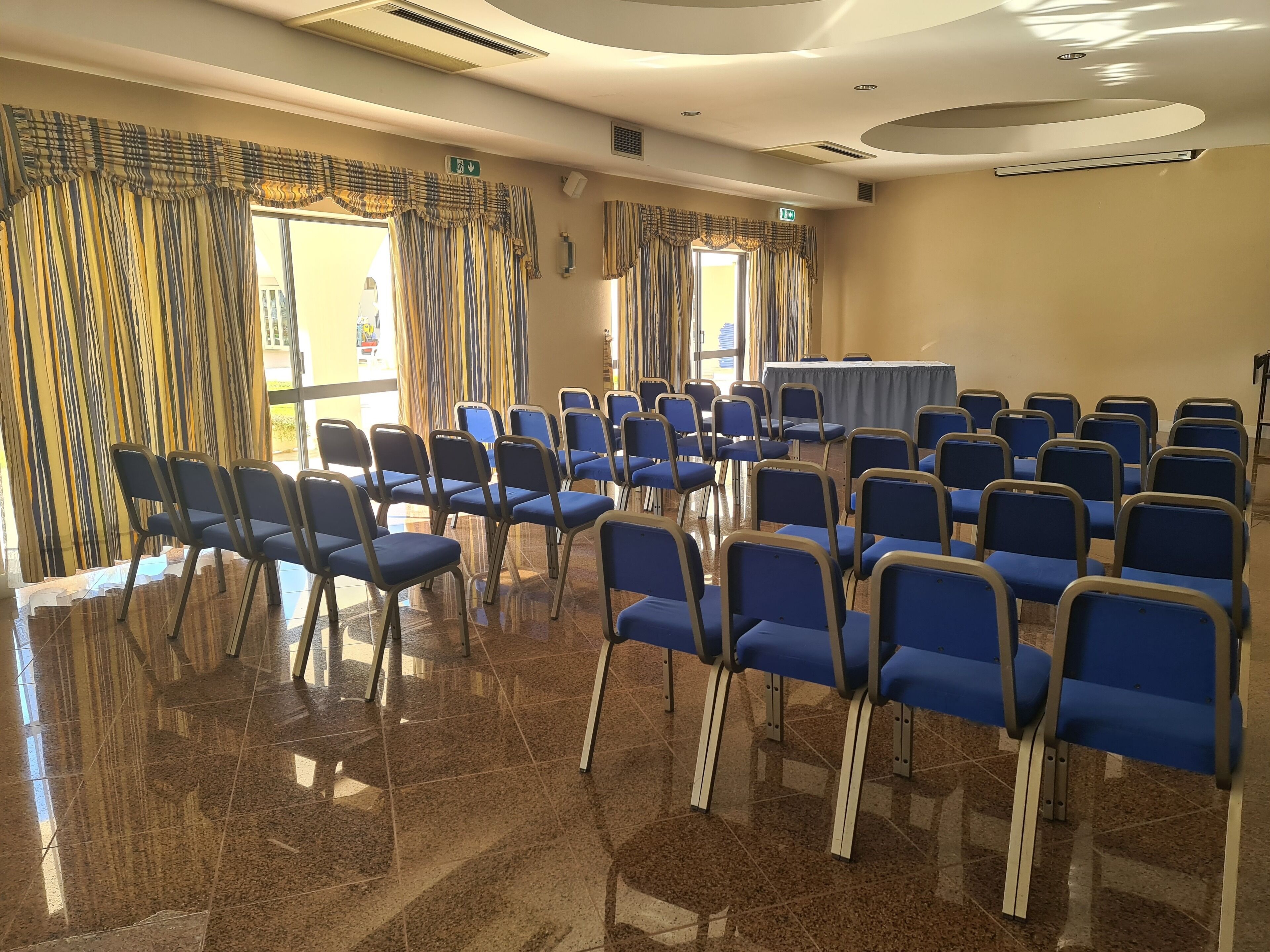 Una sala conferenze con file di sedie blu, un tavolo e ampie finestre con tende.