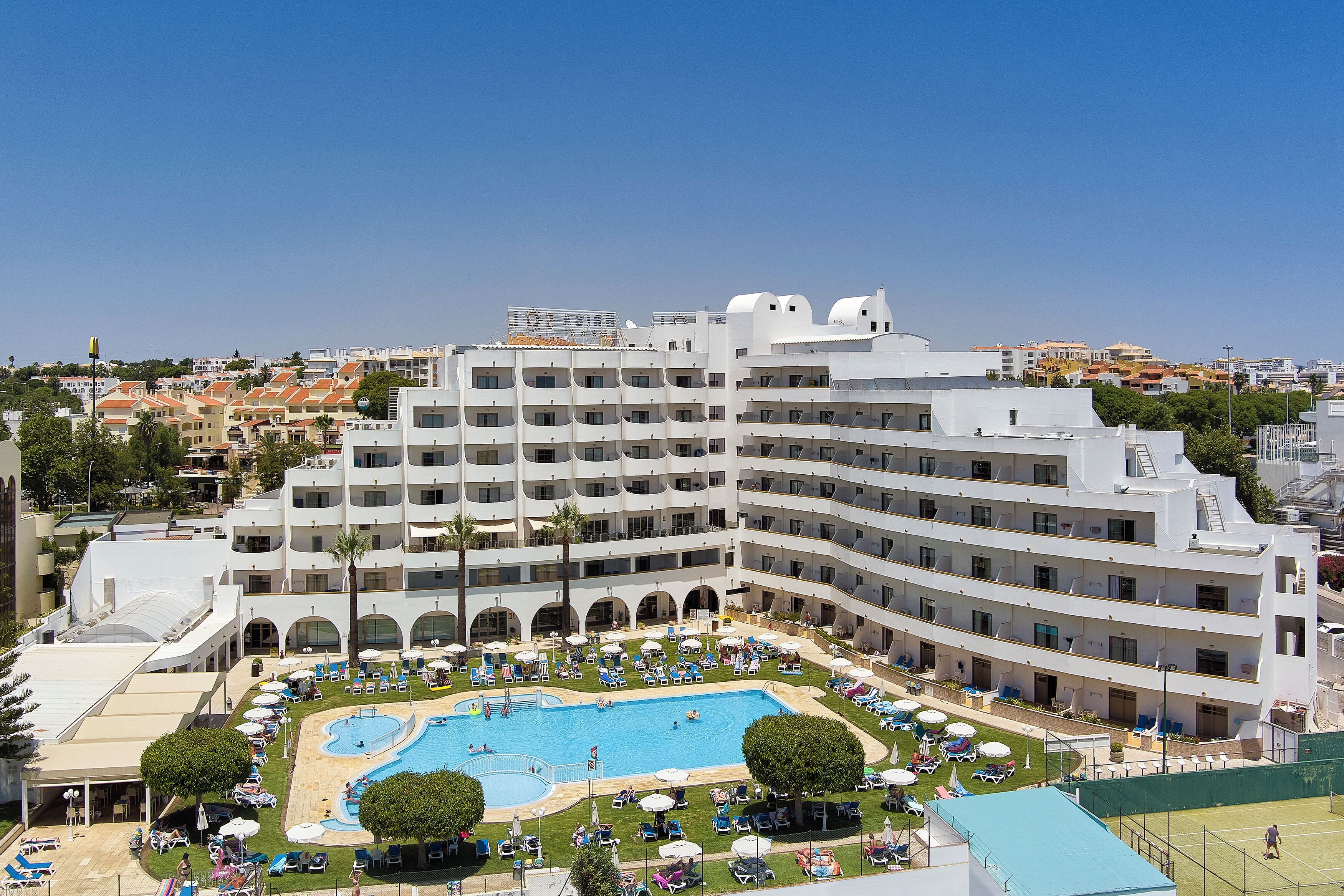 A multi-story hotel with a swimming pool, sun loungers, and a tennis court.