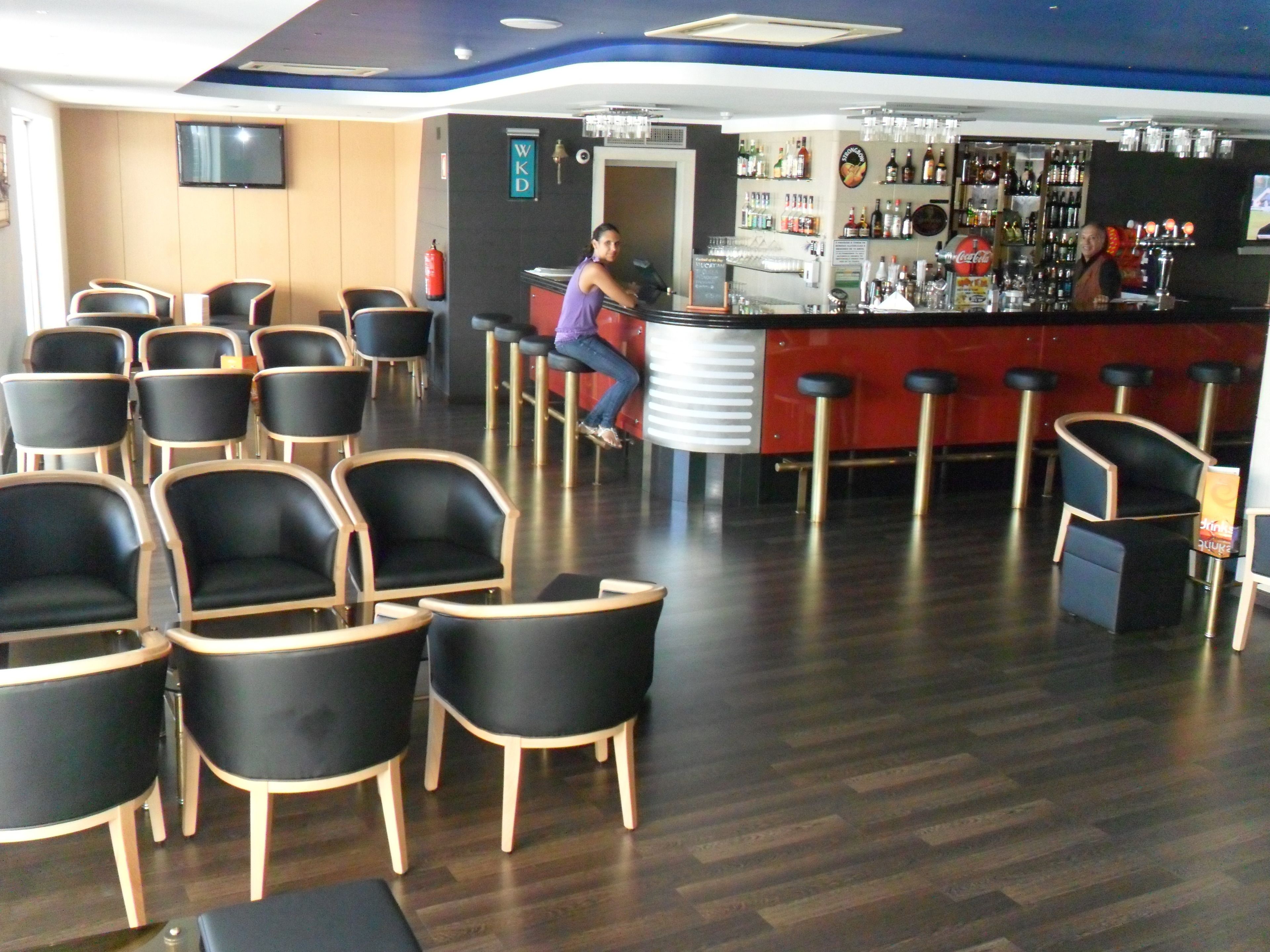 Zona de bar con sillas y taburetes negros, una barra con un bartender y estantes con botellas.
