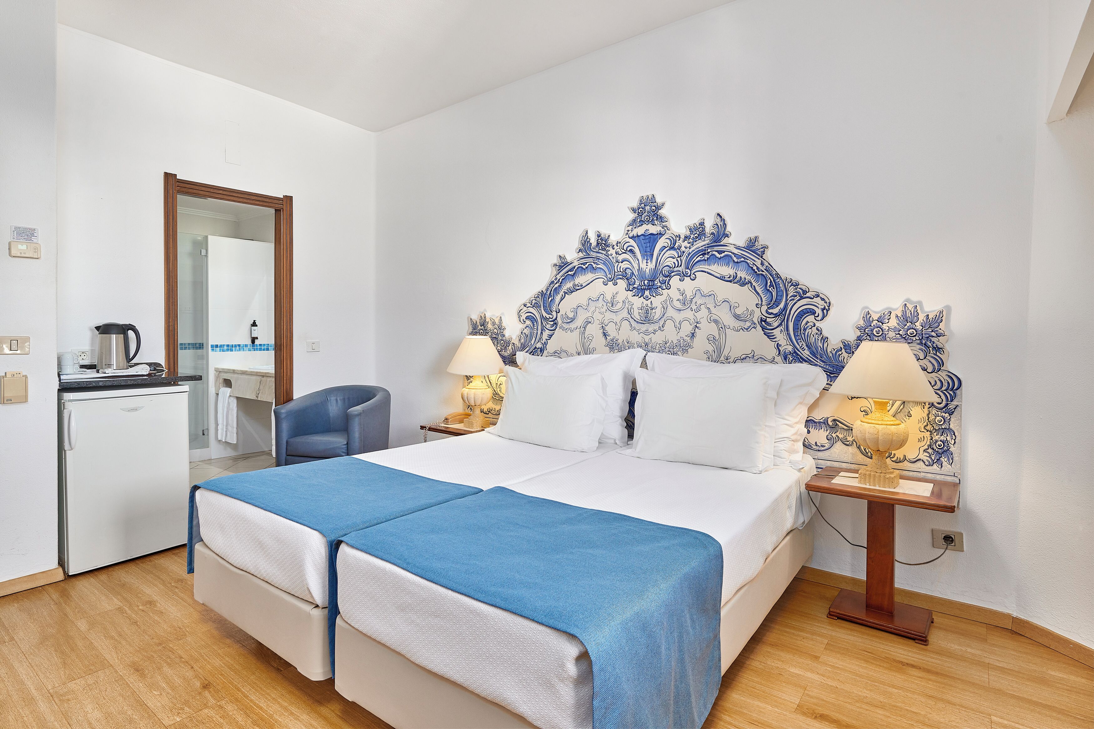 A double bed with a blue bedspread, two bedside lamps, a decorative headboard, a small desk with a kettle, and a chair.